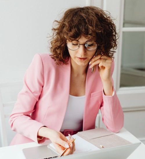 Woman in a light pink blazer on the phone and writing on a notepad