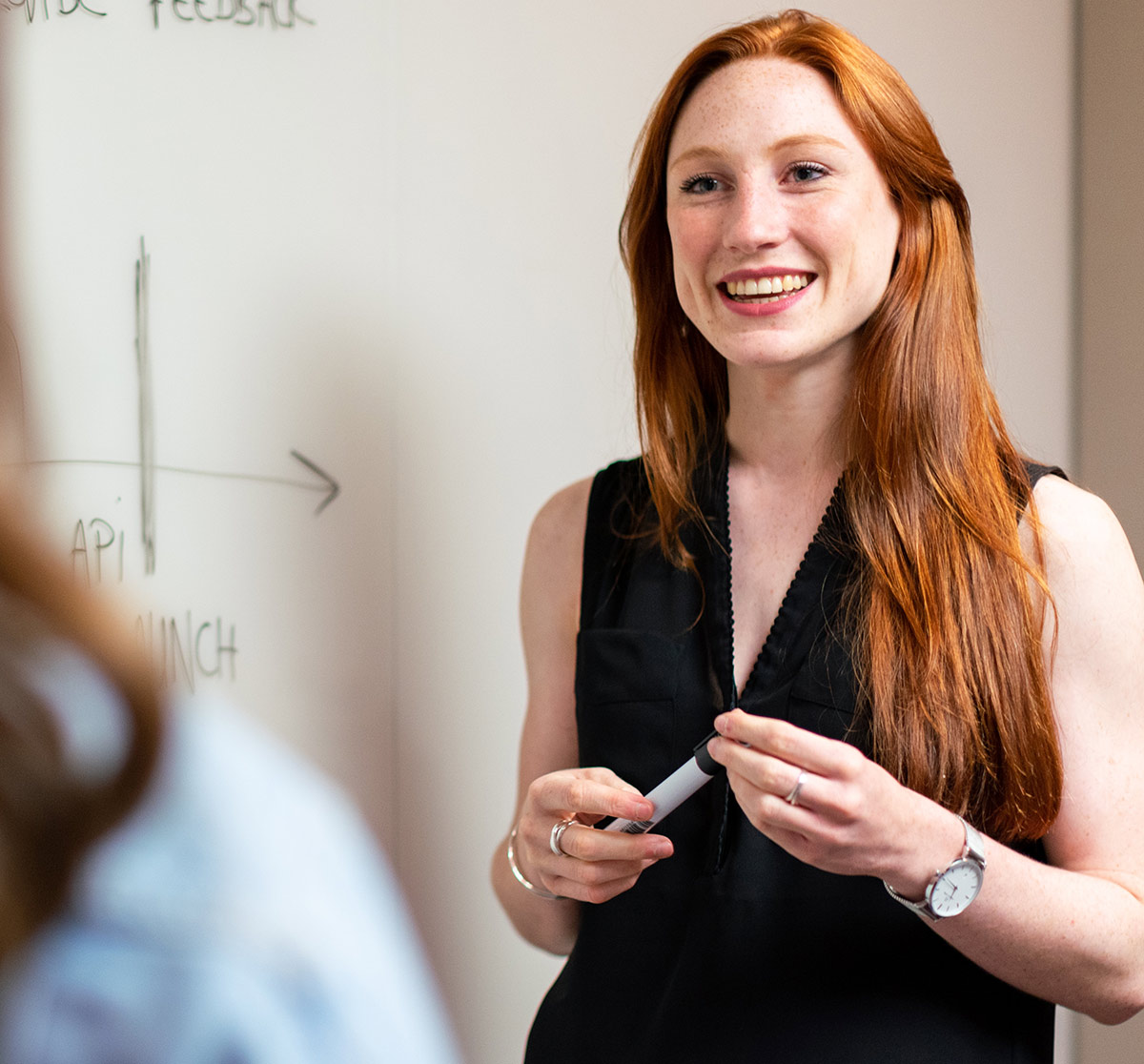 Two women face each other in front of a white board talking as one of them has a whiteboard marker in their right hand and is wearing a watch on their left hand