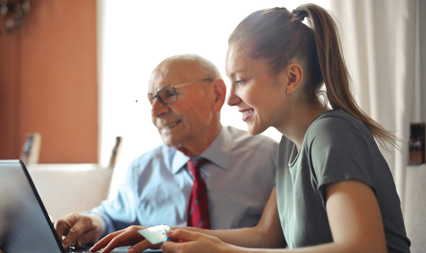 woman and elderly man smile while holding debit card and looking at a laptop screen together