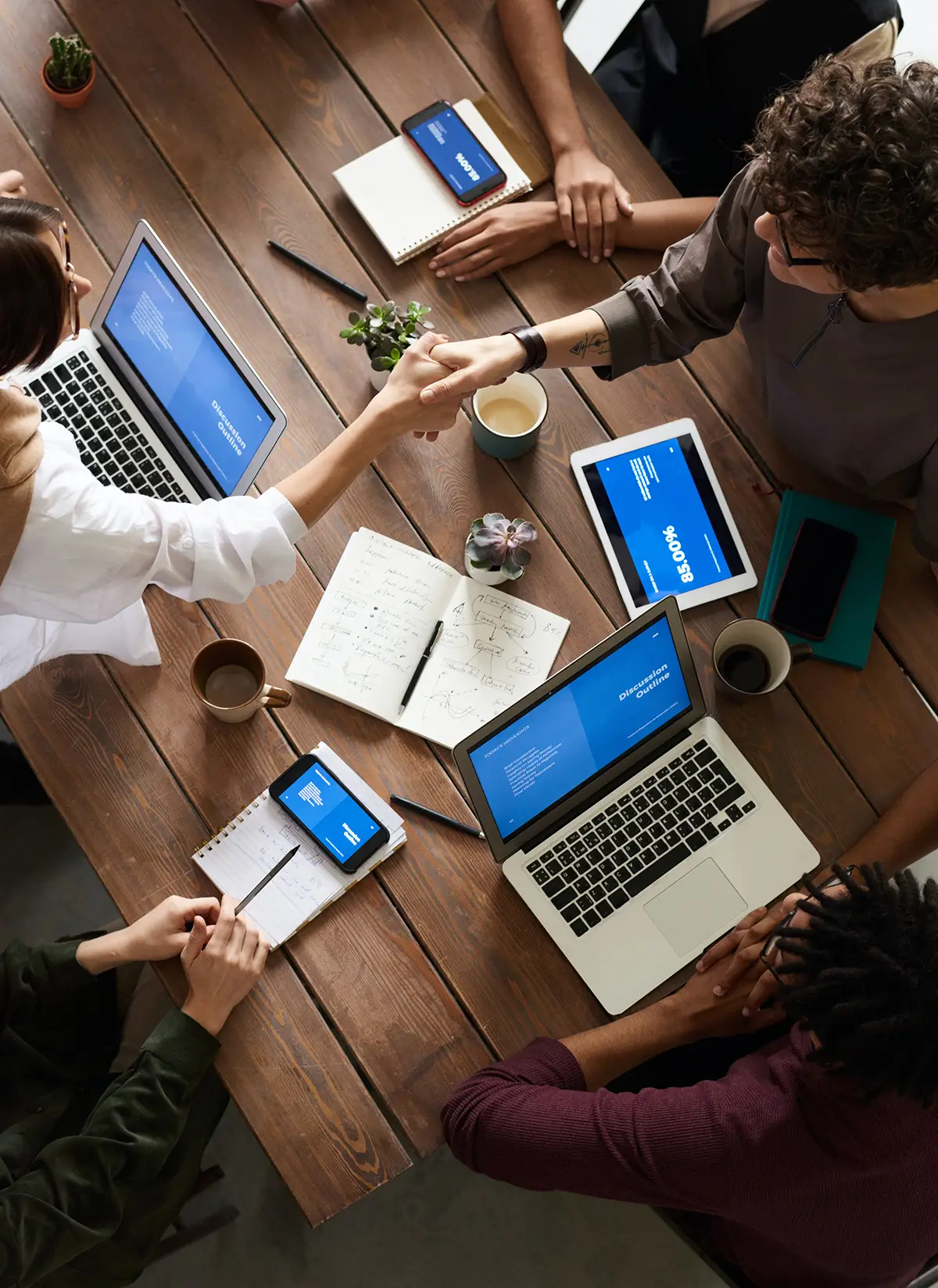 Group of individuals sitting at a table as a woman and a man are shaking hands across the table as there are laptops, a tablet, and smartphones nearby
