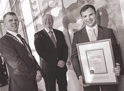 Landscape close-up sepia tone colored photograph of Conor Molloy (far right) holding an award plaque picture frame of the CPA Ireland Liam Donnelly Medal of Excellence certificate as he stands next to President of CPA Ireland, Mark Gargan (far left) and CEO of CPA Ireland, Eamonn Siggins (middle) in where all of them are smiling for a group photo