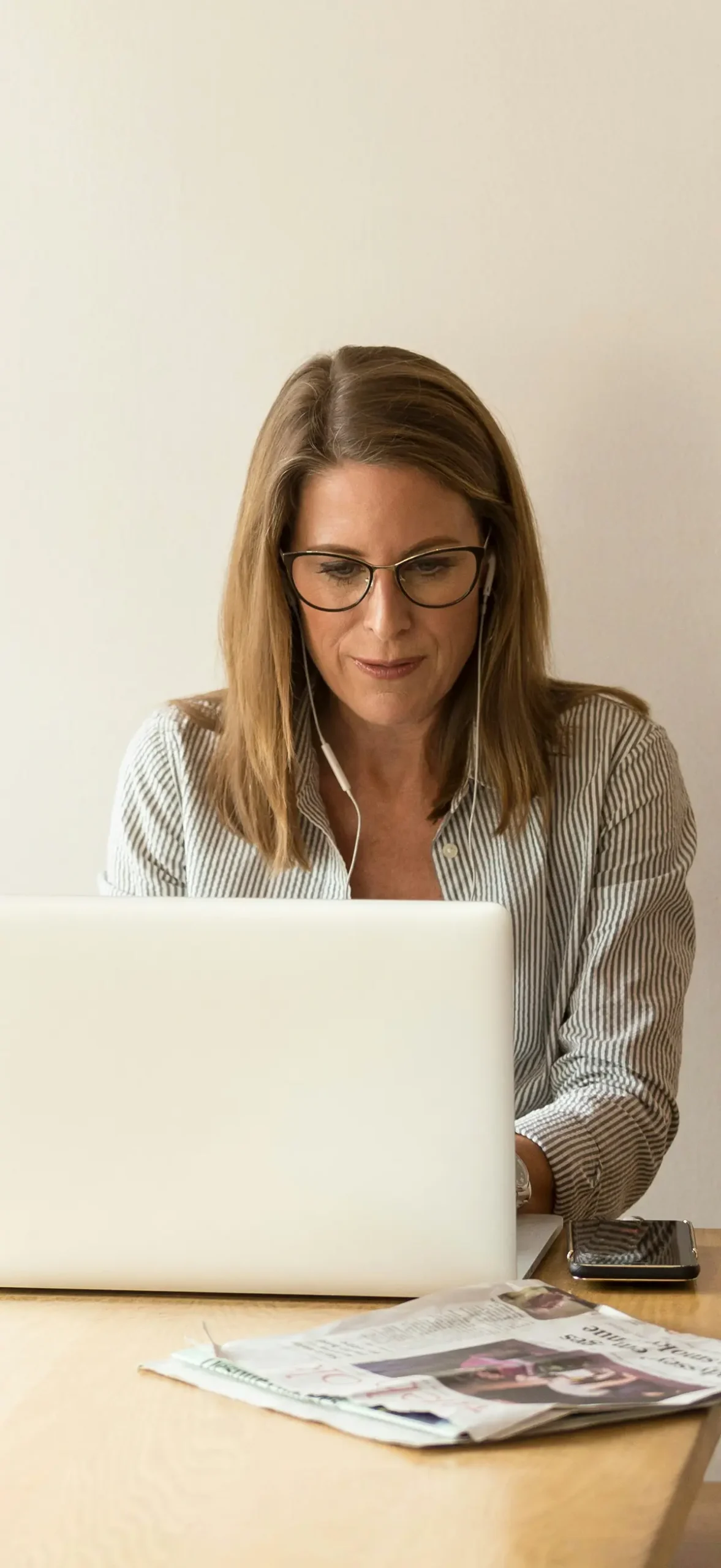 Portrait close-up photograph view of a woman listening to her wired earbuds and glancing at a laptop screen while seated down at a wooden table with a smartphone and newspaper situated nearby the laptop